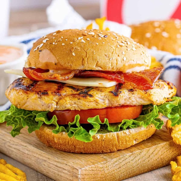 Grilled chicken sandwich with bacon, cheese, lettuce, and tomato on a wooden board with fries.