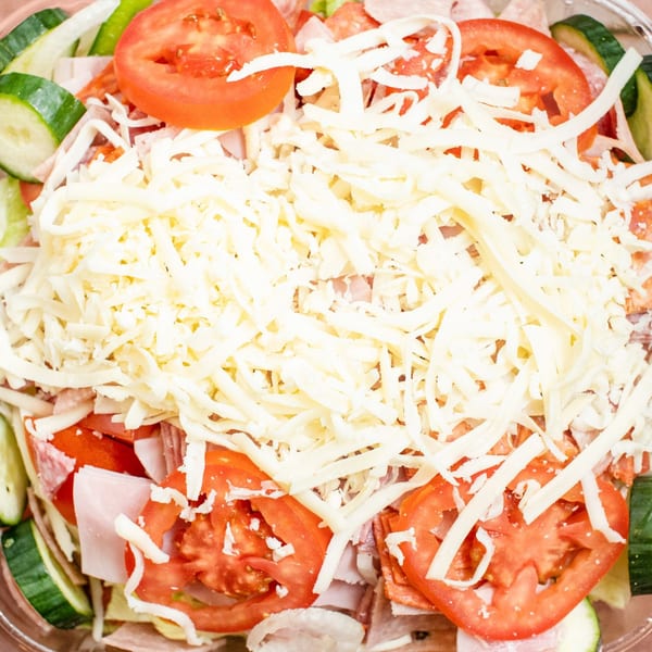 Close-up of a fresh salad topped with shredded cheese, sliced tomatoes, cucumbers, and deli meats.